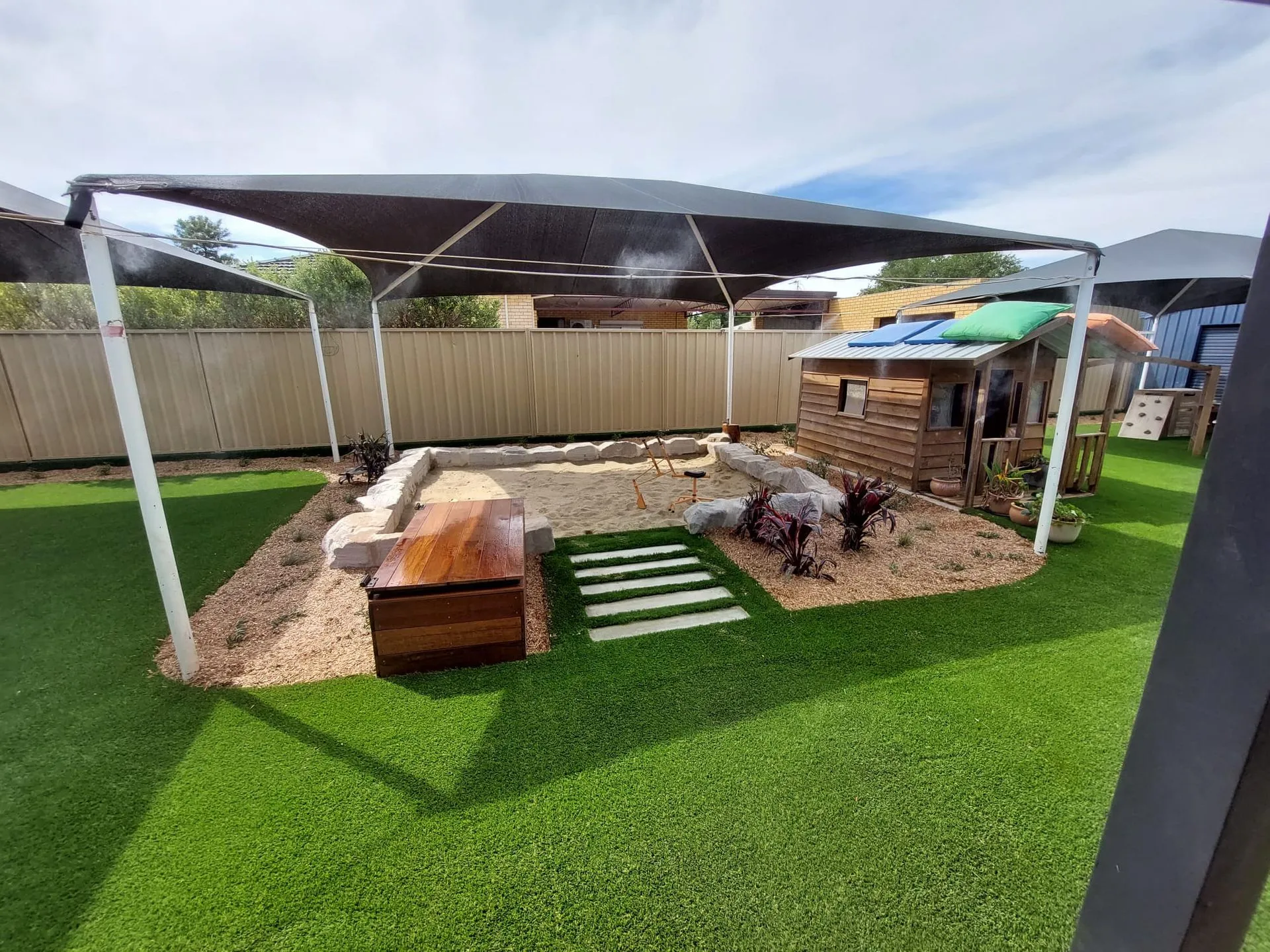 Childcare outdoor play area under shade sails with timber cubby house and sandstone seating