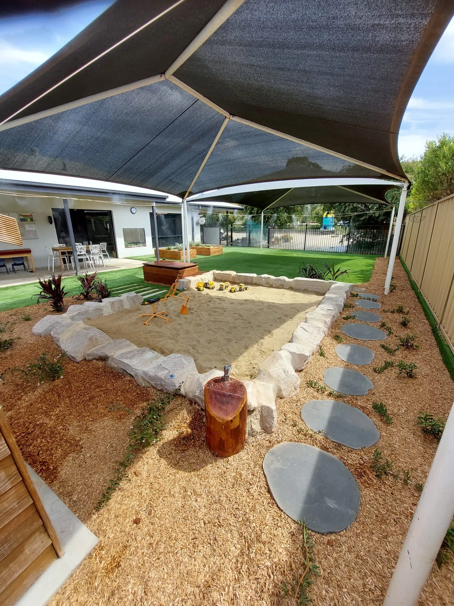 Childcare sandpit under a shade sail with stepping stones and timber stumps